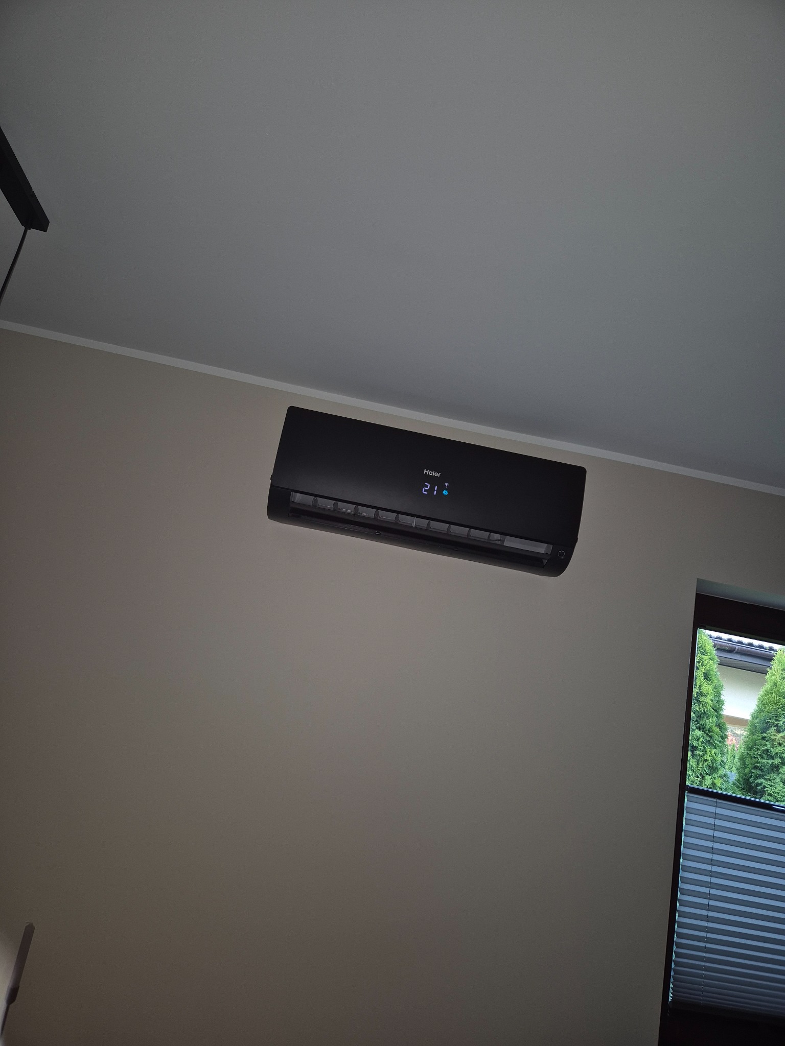 Czarny klimatyzator Haier na ścianie w pokoju, widoczne okno z żaluzjami i zielone drzewa na zewnątrz. Wyświetlacz klimatyzatora pokazuje temperaturę 21 stopni.