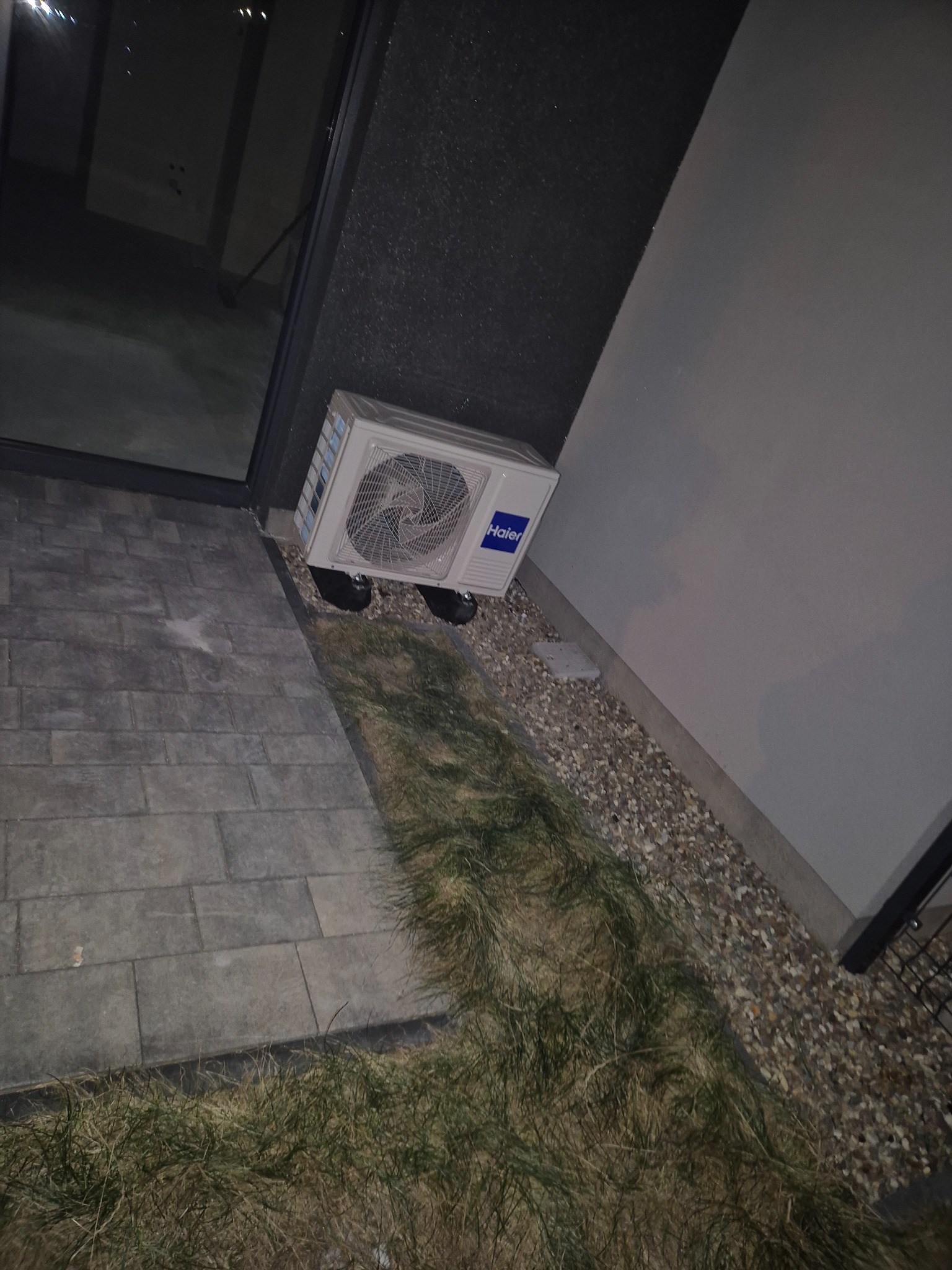 Biała jednostka zewnętrzna klimatyzacji Haier zamontowana przy ścianie budynku, widoczna z boku na tle kostki brukowej i trawy.