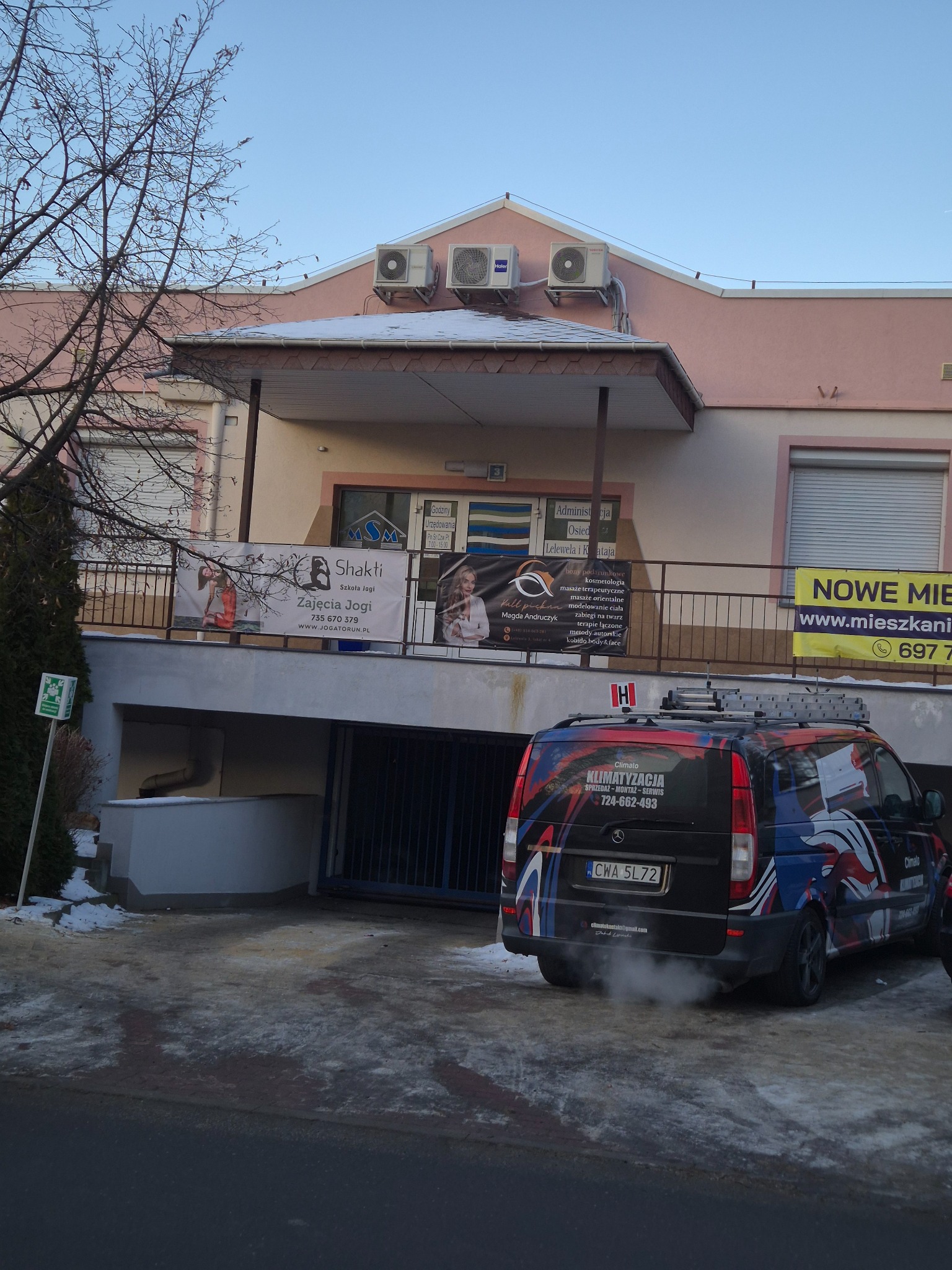 Budynek z trzema klimatyzatorami na dachu i samochodem serwisowym z napisem 'Klimatyzacja' na parkingu. Widoczne reklamy innych firm na balustradzie.