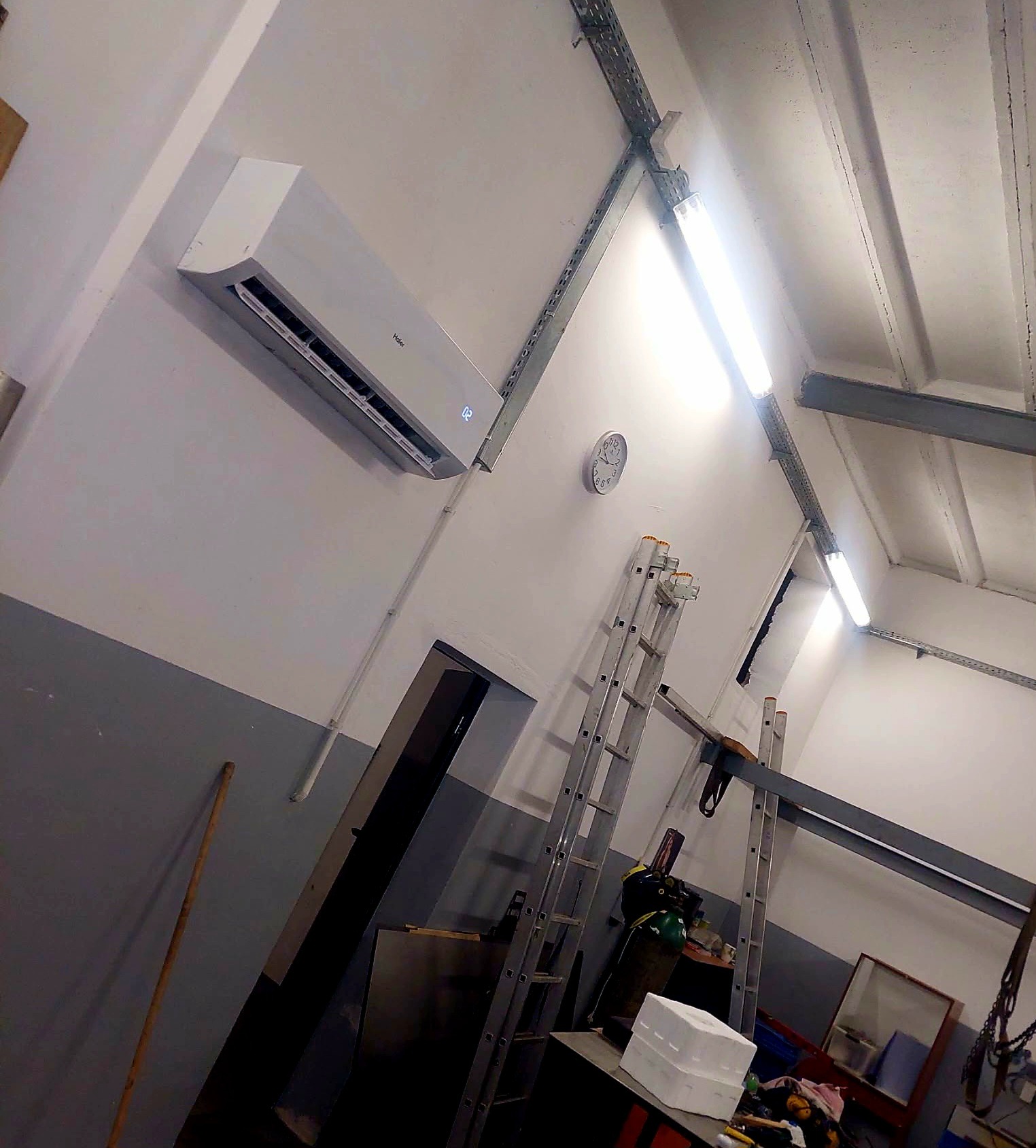 Biały klimatyzator Haier na ścianie w pomieszczeniu przemysłowym, widoczne drabiny, instalacje pod sufitem i zegar. Ujęcie z dołu.