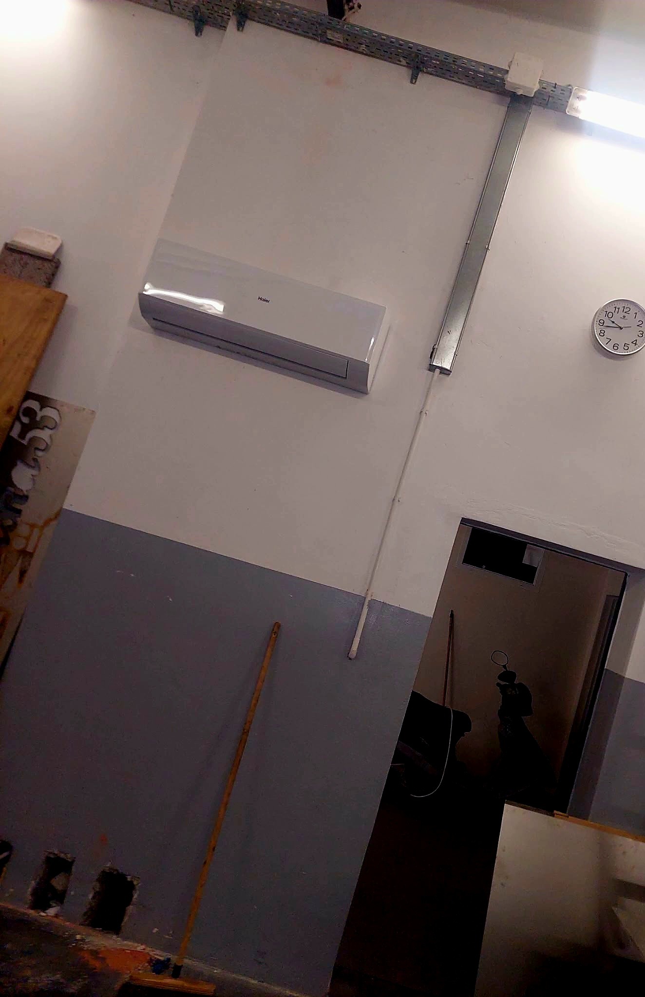 Biały klimatyzator Haier na ścianie nad szarą lamperią, obok zegar analogowy i instalacja elektryczna. Widoczny fragment pomieszczenia w trakcie prac wykończeniowych.