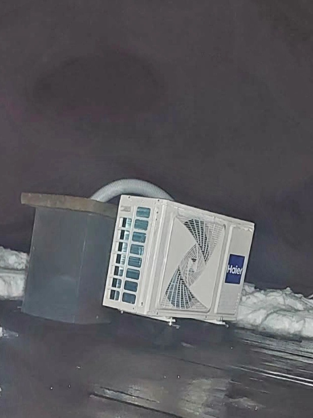 Biały klimatyzator Haier z podłączonym przewodem, zamontowany na zewnątrz budynku, widoczny w zimowej scenerii z resztkami śniegu.