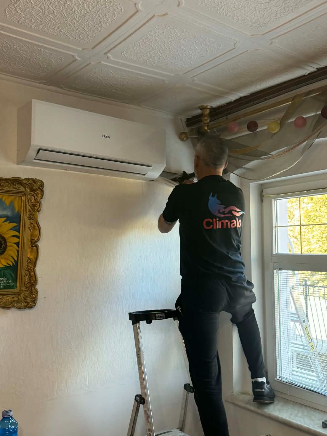 Montaż klimatyzacji Haier w domu: instalator na drabinie, widoczny klimatyzator na ścianie, dekoracyjne wykończenie sufitu i obraz w złotej ramie.