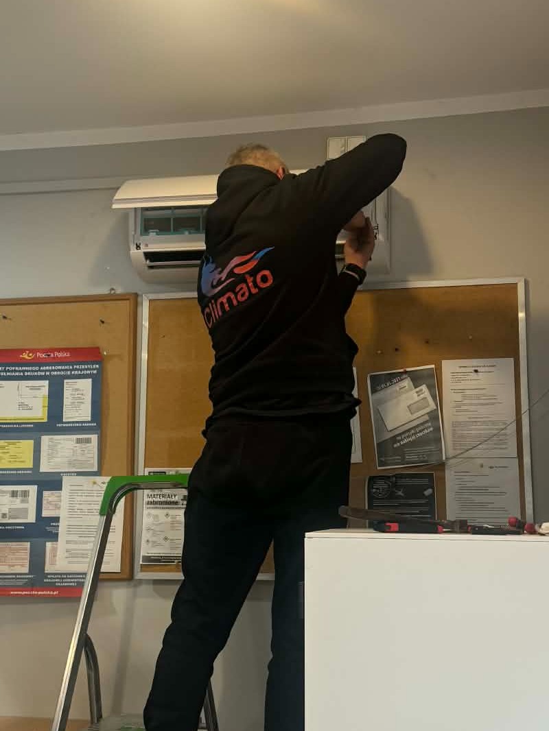 Mężczyzna w bluzie z logo 'Climato' na drabinie, montuje klimatyzator ścienny w pomieszczeniu biurowym. Na ścianie tablice informacyjne i narzędzia.