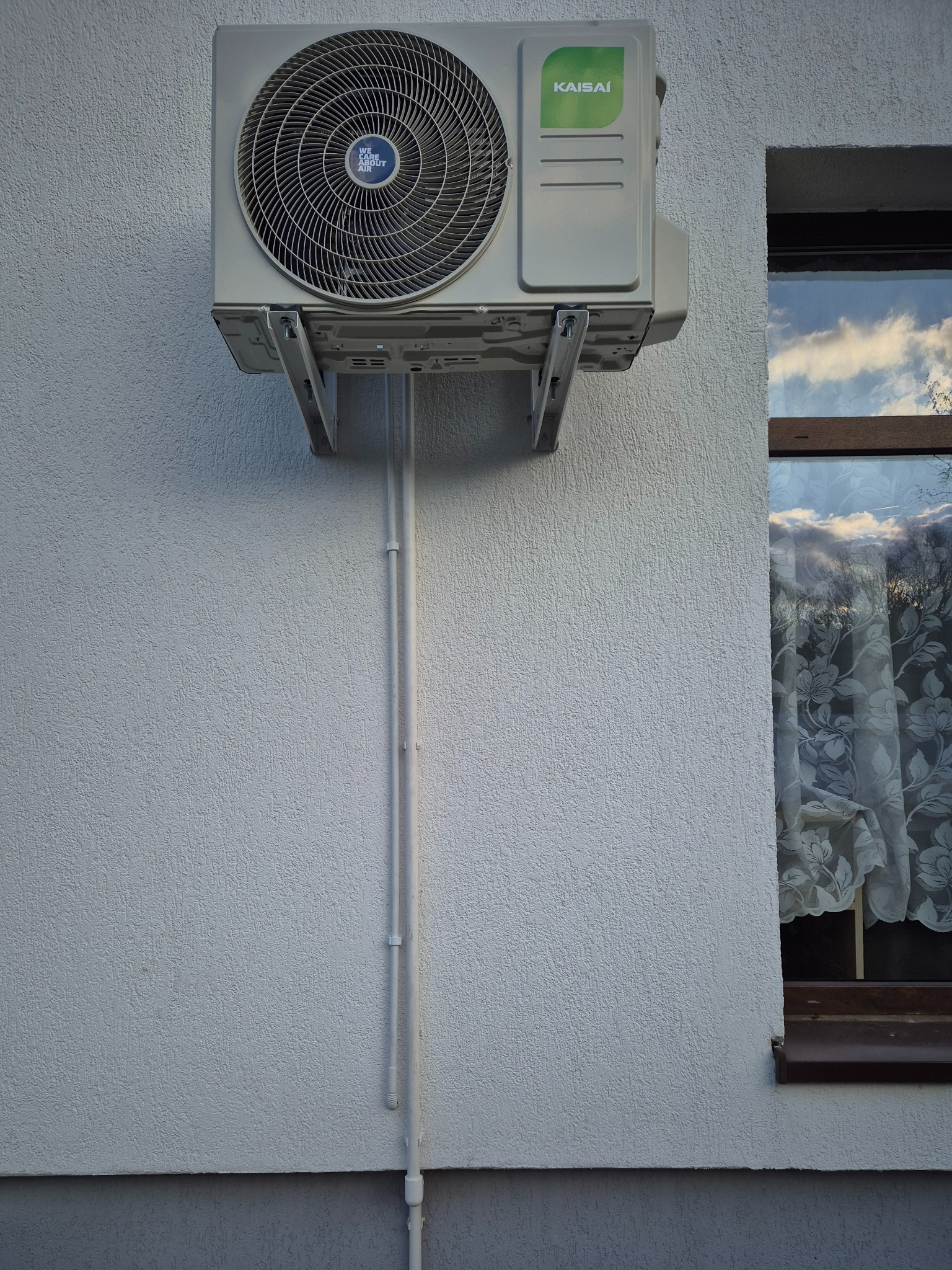 Zamontowana jednostka zewnętrzna klimatyzacji Kaisai na elewacji budynku, widoczne rury instalacyjne i okno z firanką. Jasna elewacja, dzień.