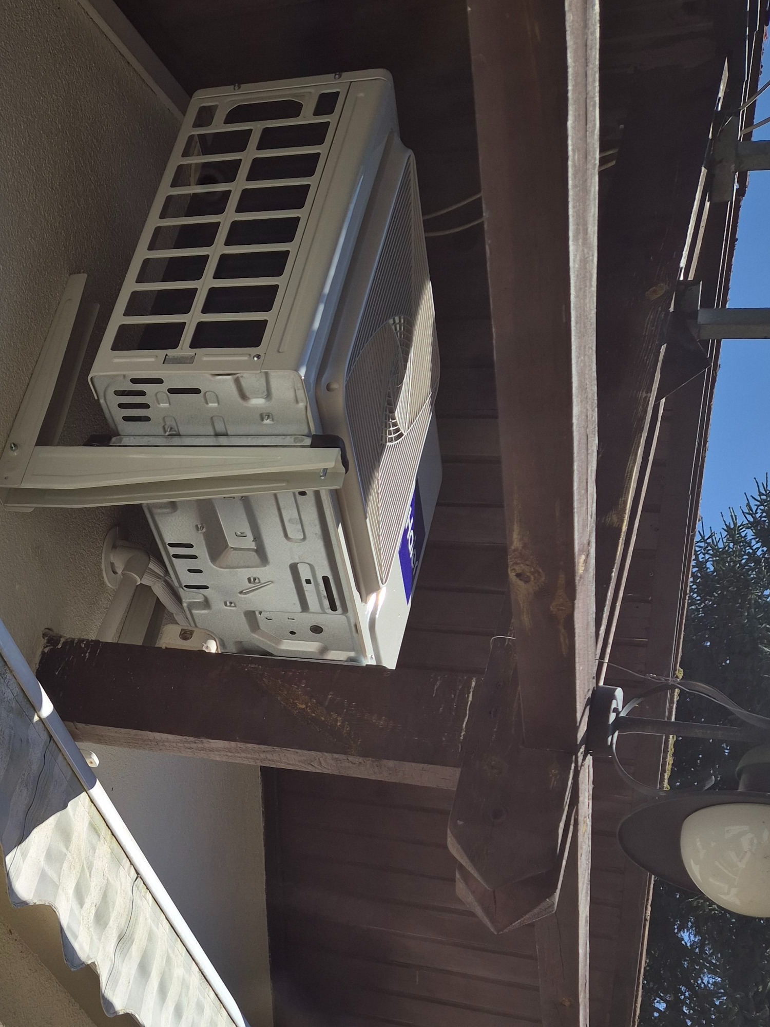 Zamontowana jednostka zewnętrzna klimatyzacji marki Haier na ścianie budynku, widoczna spod drewnianej konstrukcji z oświetleniem. Błękitne niebo w tle.