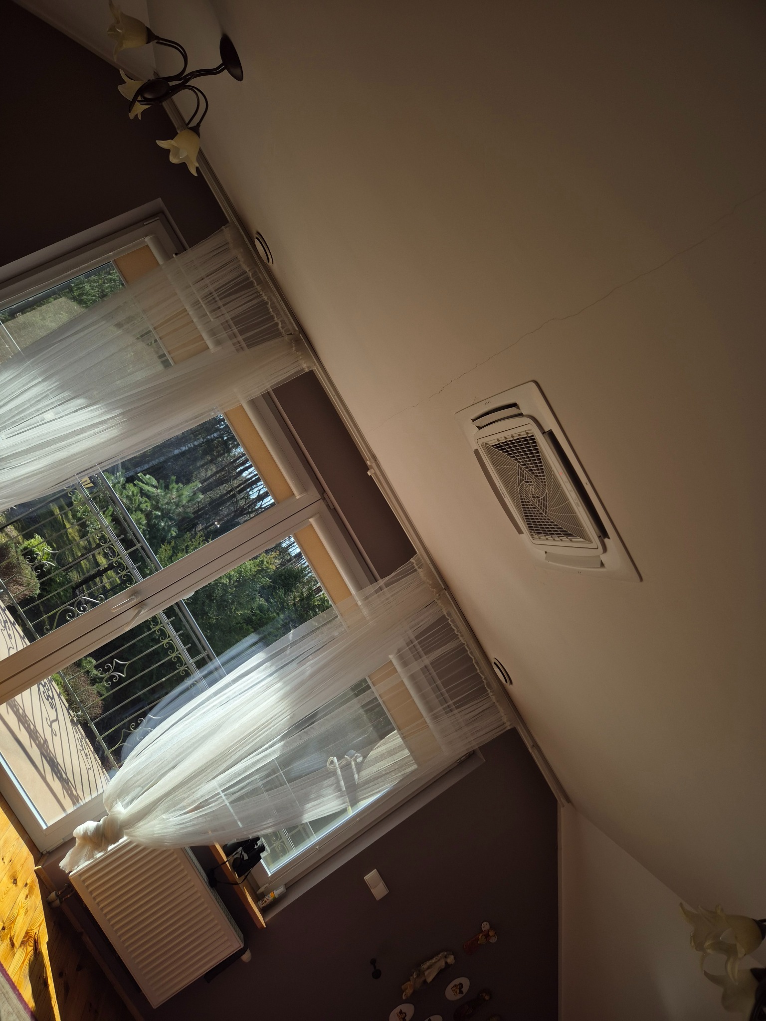 Kasetonowa klimatyzacja sufitowa w jasnym wnętrzu, widok z dołu. Okno z firanką i grzejnik w tle. Ujęcie z perspektywy żabiej.