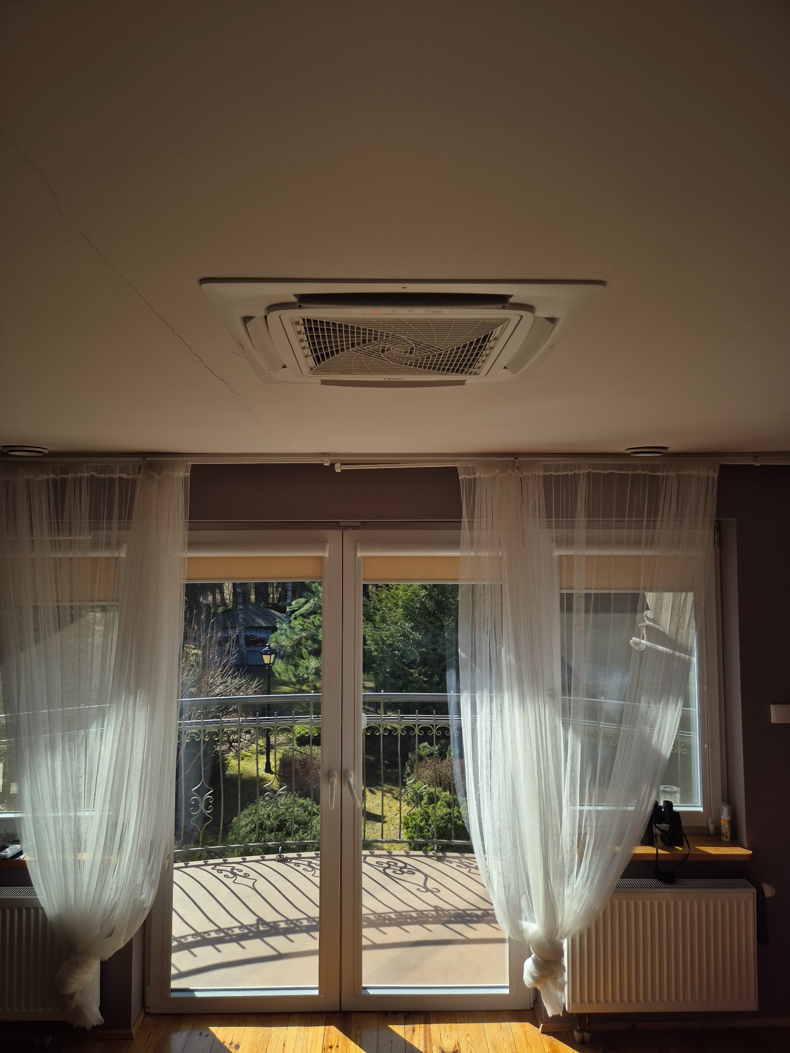 Kasetonowa klimatyzacja sufitowa w jasnym wnętrzu z balkonem i firankami. Widok na zieleń za oknem. Promienie słoneczne podkreślają detale instalacji.