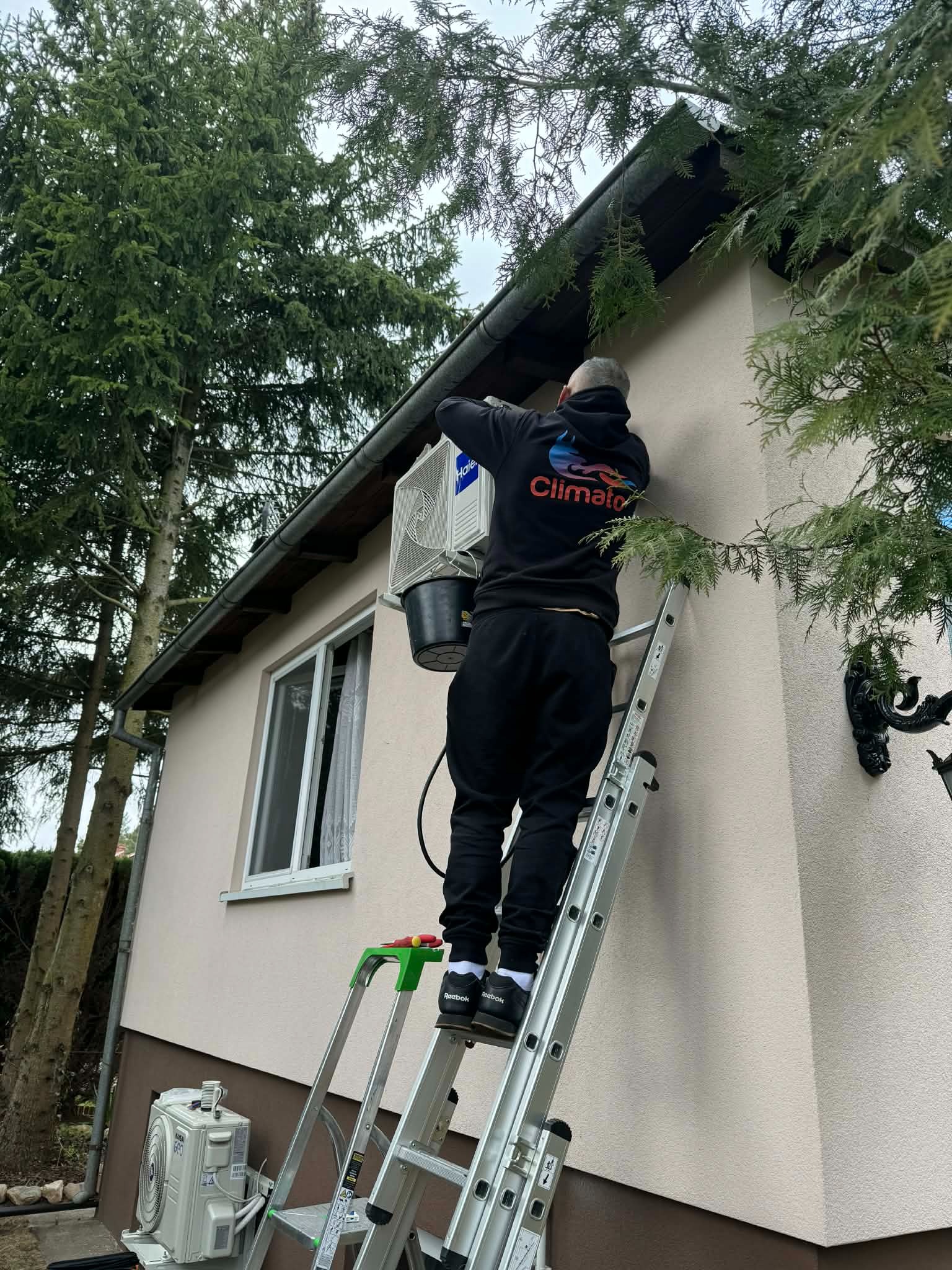Mężczyzna na drabinie montuje klimatyzator na elewacji domu. Widoczny czarny kabel i wiadro. Na dole drugi klimatyzator. Ubrany w czarną bluzę z napisem 'Climato'.