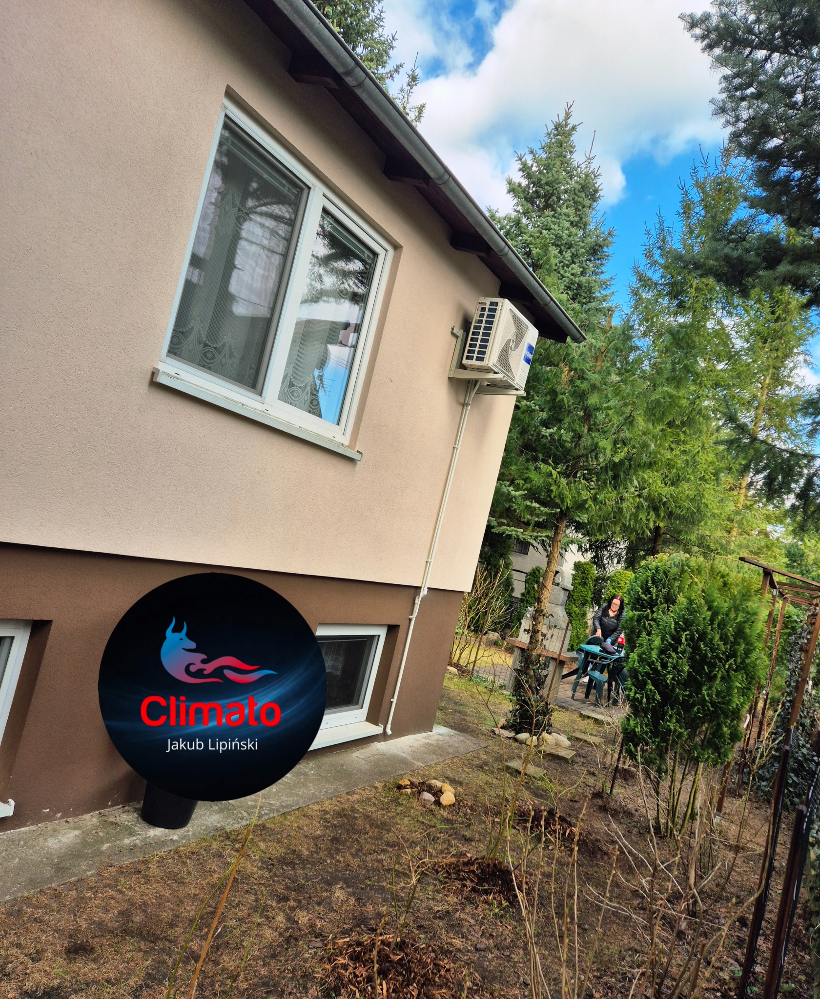 Klimatyzator na elewacji domu z widocznymi rurami instalacyjnymi. W tle zieleń ogrodu i osoba siedząca przy stole. Na pierwszym planie logo firmy.