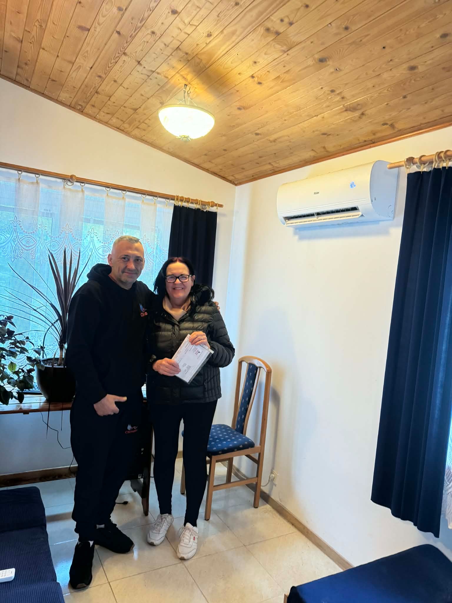 Klimatyzator Haier zamontowany w pokoju z drewnianym sufitem. Dwie osoby stoją obok, kobieta trzyma dokumenty. Widoczne zasłony i roślina doniczkowa.