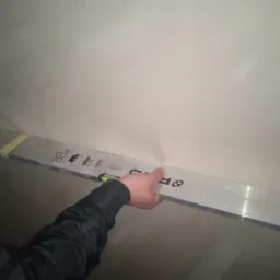 Sprawdzanie poziomu tynku maszynowego na ścianie za pomocą poziomicy aluminiowej, widoczny fragment dłoni w czarnej kurtce.