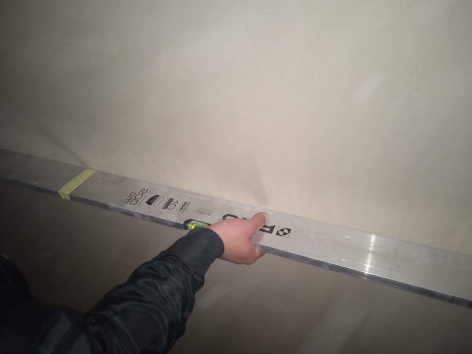 Sprawdzanie poziomu tynku maszynowego na ścianie za pomocą poziomicy aluminiowej, widoczny fragment dłoni w czarnej kurtce.