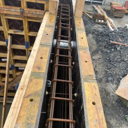 Adam Sobolewski - Szczegółowe ujęcie z góry na szalunek zbrojeniowy z ułożonymi prętami zbrojeniowymi wewnątrz, przygotowany do zalania betonem, widoczne drewniane elementy szalunku i rury wewnątrz konstrukcji.