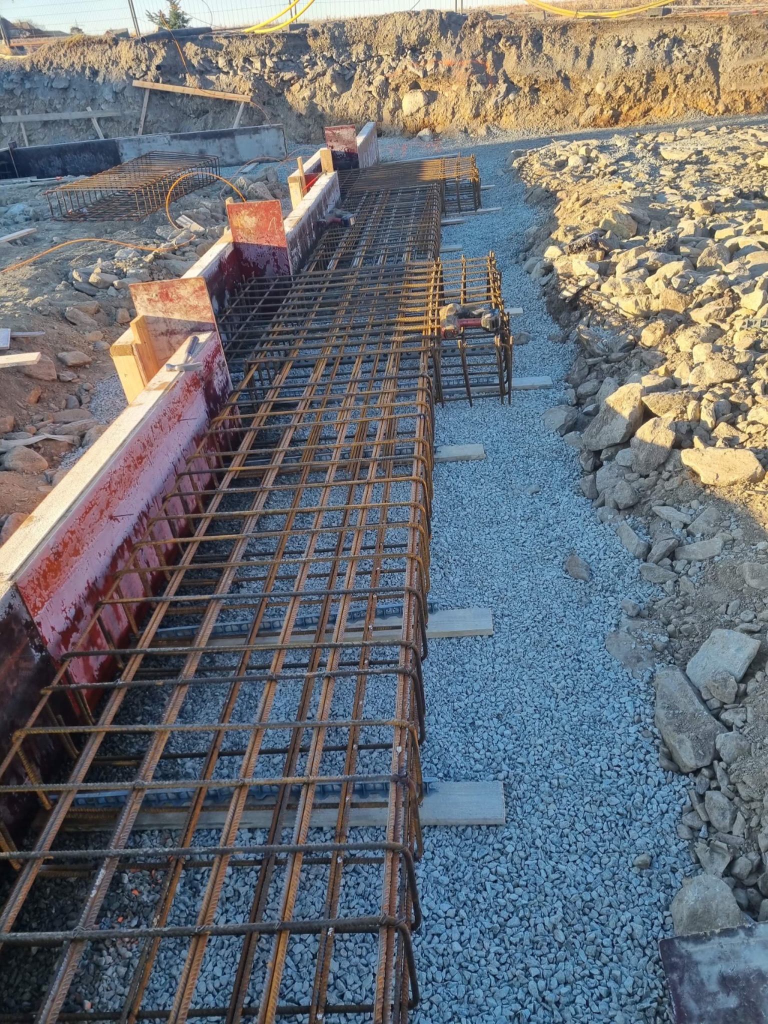 Przygotowanie zbrojenia fundamentów w szalunkach, widoczne siatki zbrojeniowe, podsypka żwirowa i drewniane elementy konstrukcyjne.
