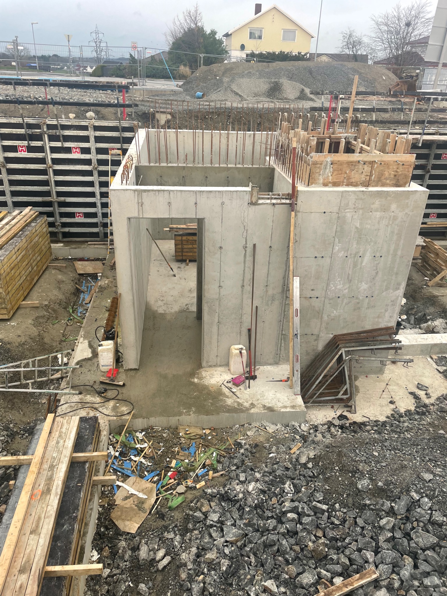 Surowa betonowa konstrukcja z otworami, prawdopodobnie piwnica lub fundament budynku, z wystającymi prętami zbrojeniowymi i drewnianymi szalunkami w tle, otoczona gruzem i materiałami budowlanymi.