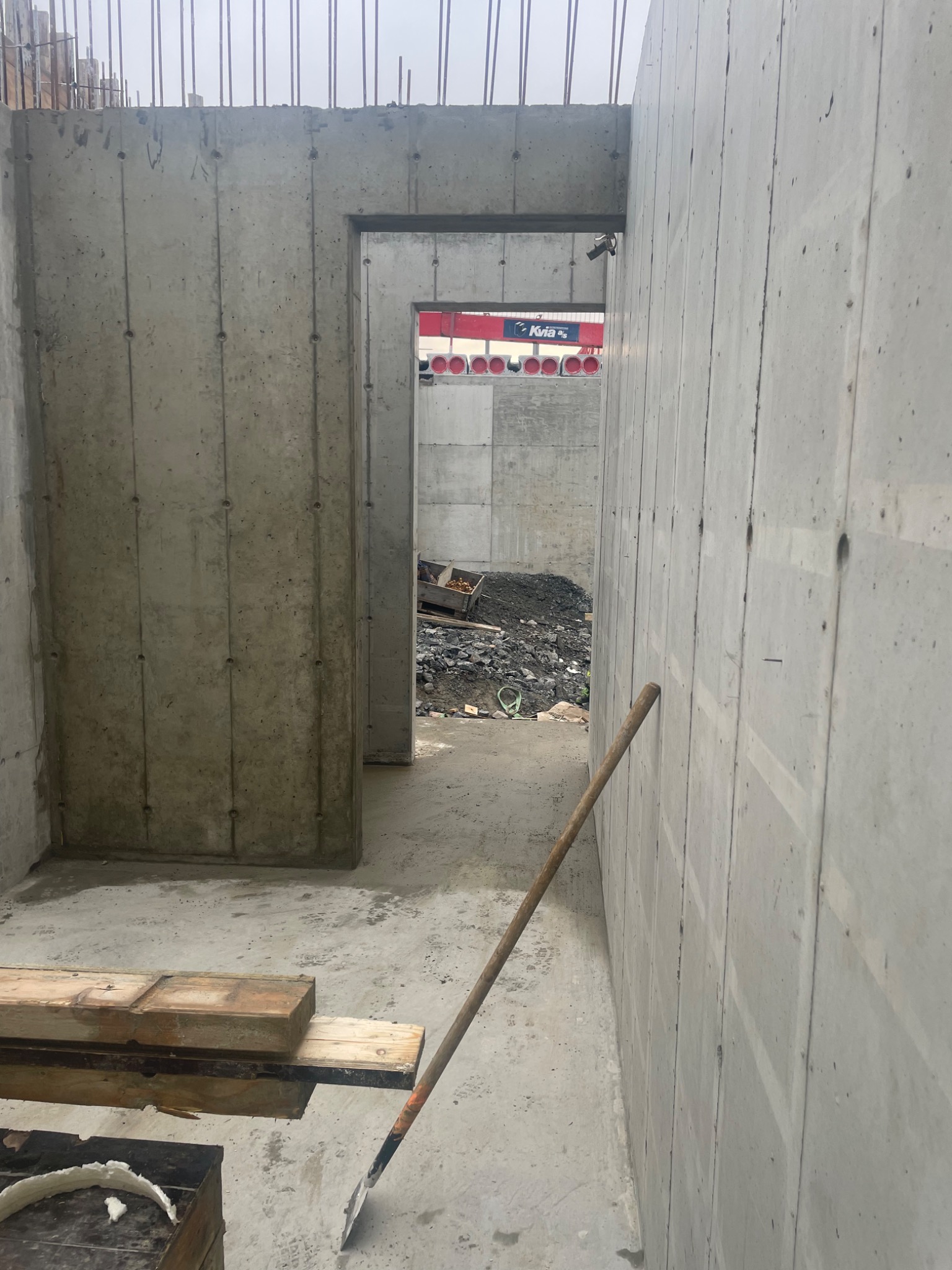 Wnętrze budynku w trakcie budowy z żelbetu, widoczne ściany z betonu, szpadel oparty o ścianę, w tle gruz i ciężarówka z dźwigiem.