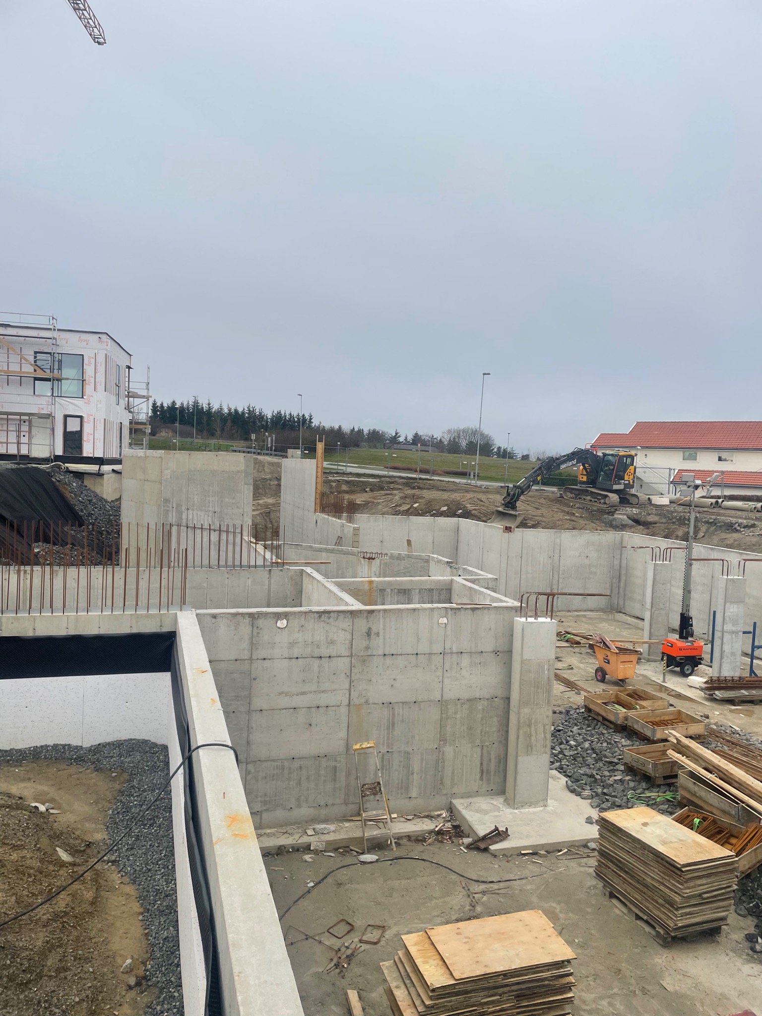 Wysoki kąt widzenia na plac budowy z wykopami fundamentów, gdzie widoczne są betonowe ściany szalunkowe z wystającymi prętami zbrojeniowymi, koparka na dalszym planie i materiały budowlane...