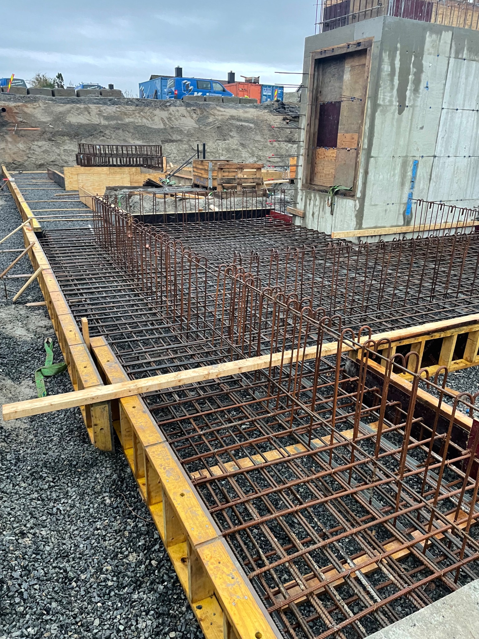 Siatka zbrojeniowa ułożona w szalunku fundamentowym, widoczne drewniane elementy konstrukcji i kamieniste podłoże, w tle ziemny nasyp i budowlane kontenery.