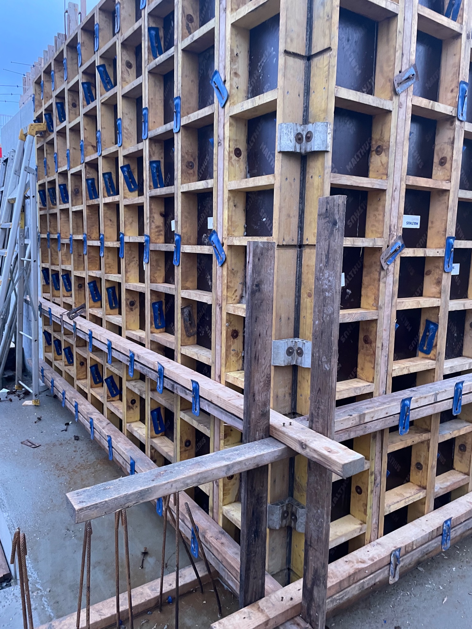 Szczegółowe ujęcie szalunku z desek i płyt z widocznymi niebieskimi zaciskami, przygotowanego do zalewania betonem, z wystającymi prętami zbrojeniowymi w dole kadru.