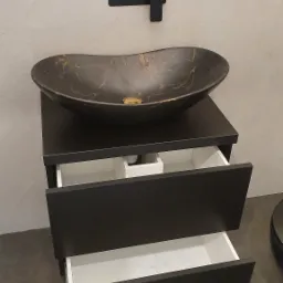 Czarna szafka łazienkowa z dwoma wysuniętymi szufladami, na której stoi owalna umywalka z czarnego marmuru ze złotymi żyłkami, czarna bateria ścienna i gniazdko elektryczne w tle.