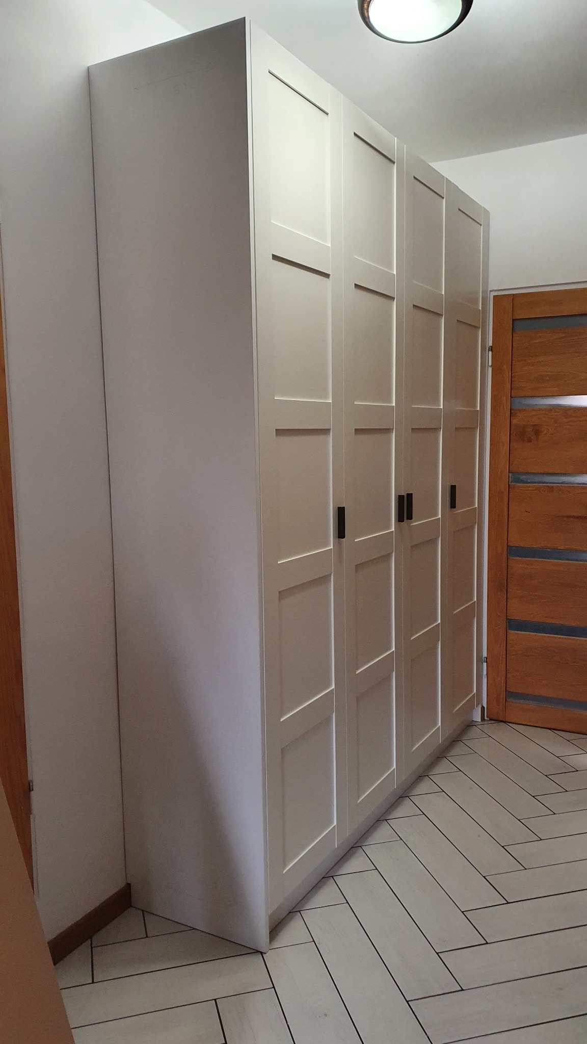 Biała szafa wnękowa z czterema skrzydłami drzwiowymi, z czarnymi uchwytami, dopasowana do skosu ściany w mieszkaniu w Józefinie.