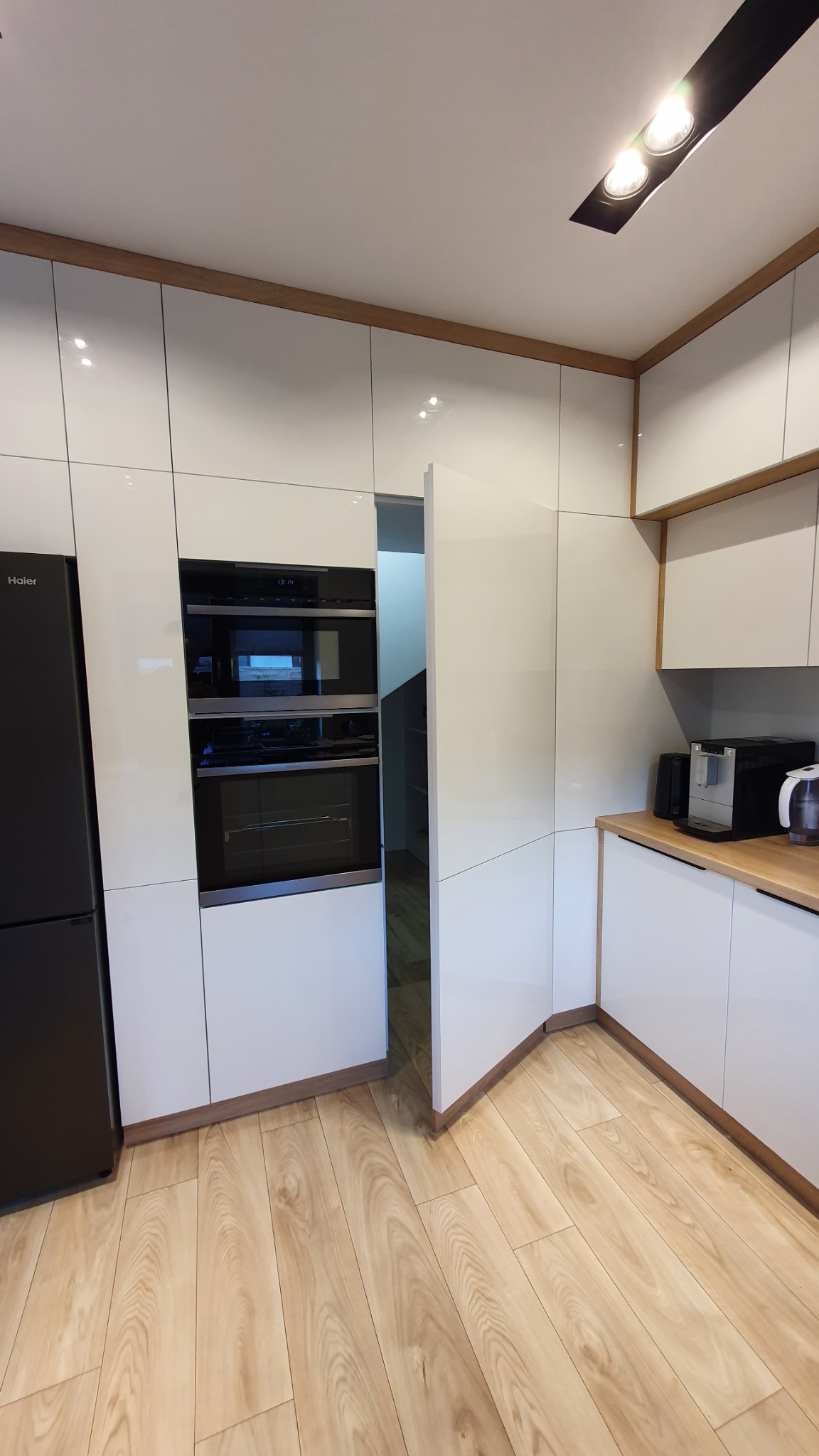 Nowoczesna kuchnia z białymi, lakierowanymi frontami szafek, drewnianymi akcentami i ukrytymi drzwiami w zabudowie, z widocznym piekarnikiem i lodówką marki Haier.