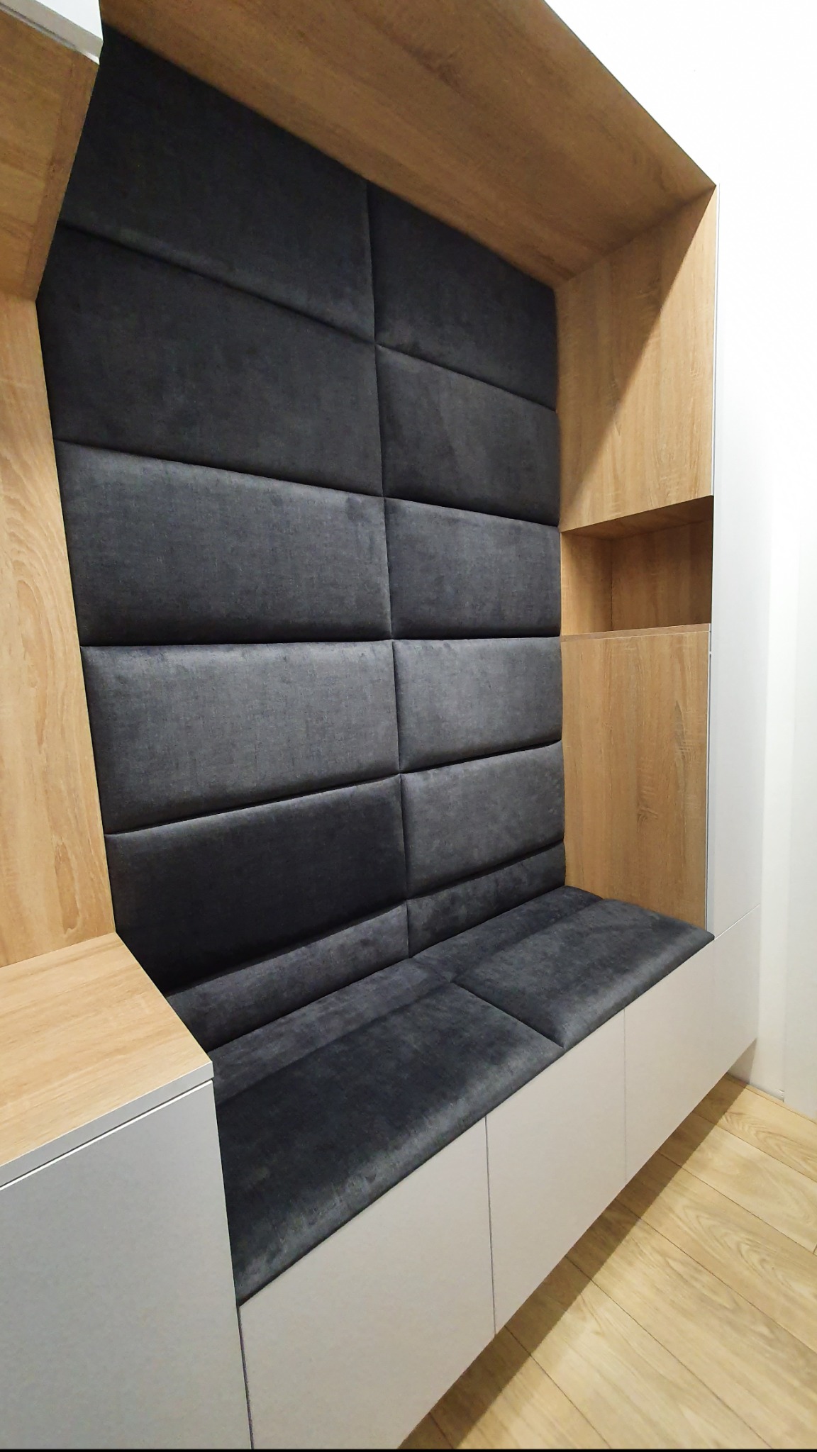 Wnęka z tapicerowanym siedziskiem i ścianą z paneli w odcieniach szarości, obudowana drewnianą konstrukcją z półkami, na podłodze panele.