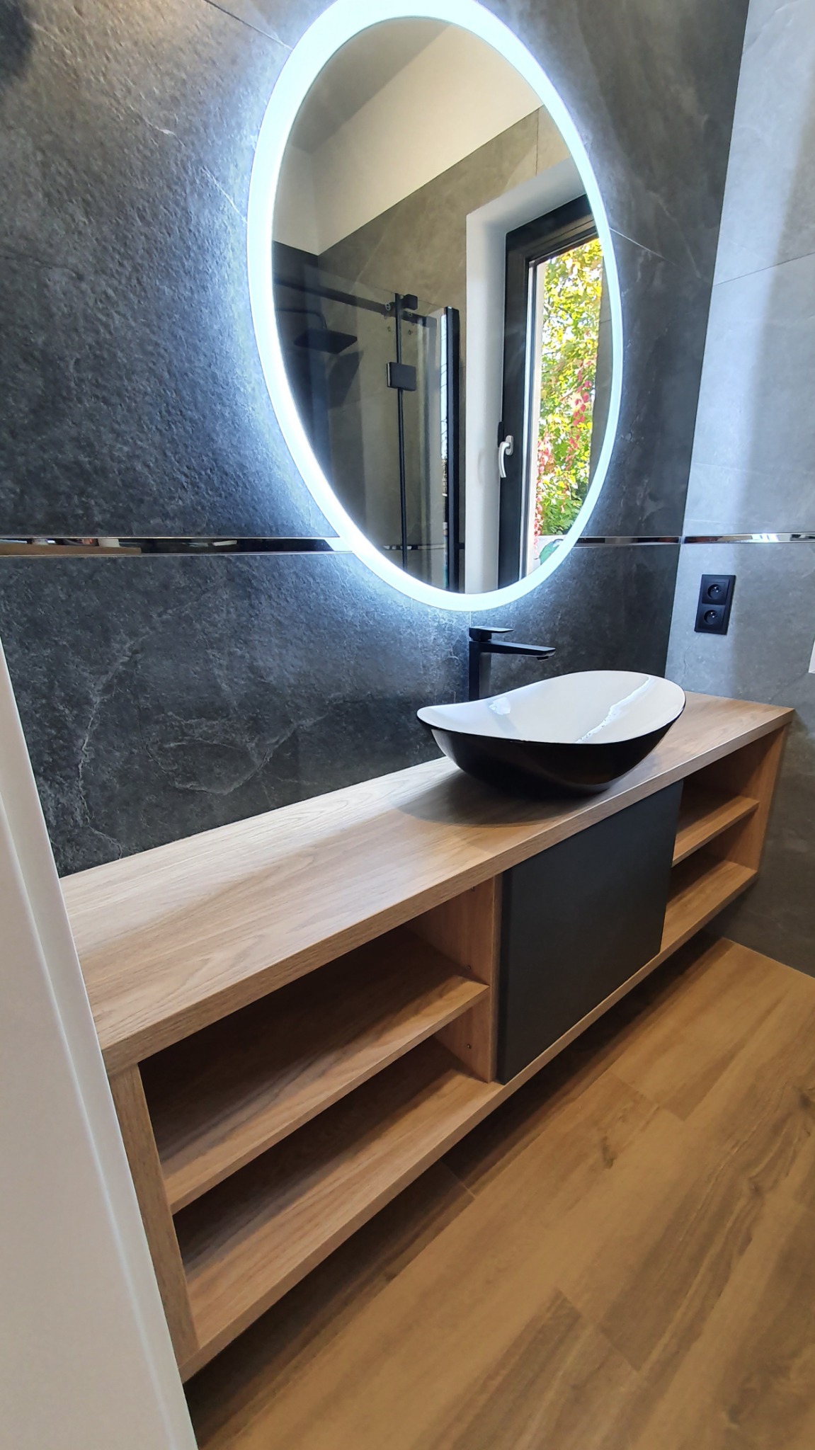 Nowoczesna łazienka z podświetlanym, okrągłym lustrem nad drewnianą szafką z półkami i czarnym elementem, na której stoi designerska umywalka w kolorze czarno-białym z czarną baterią.