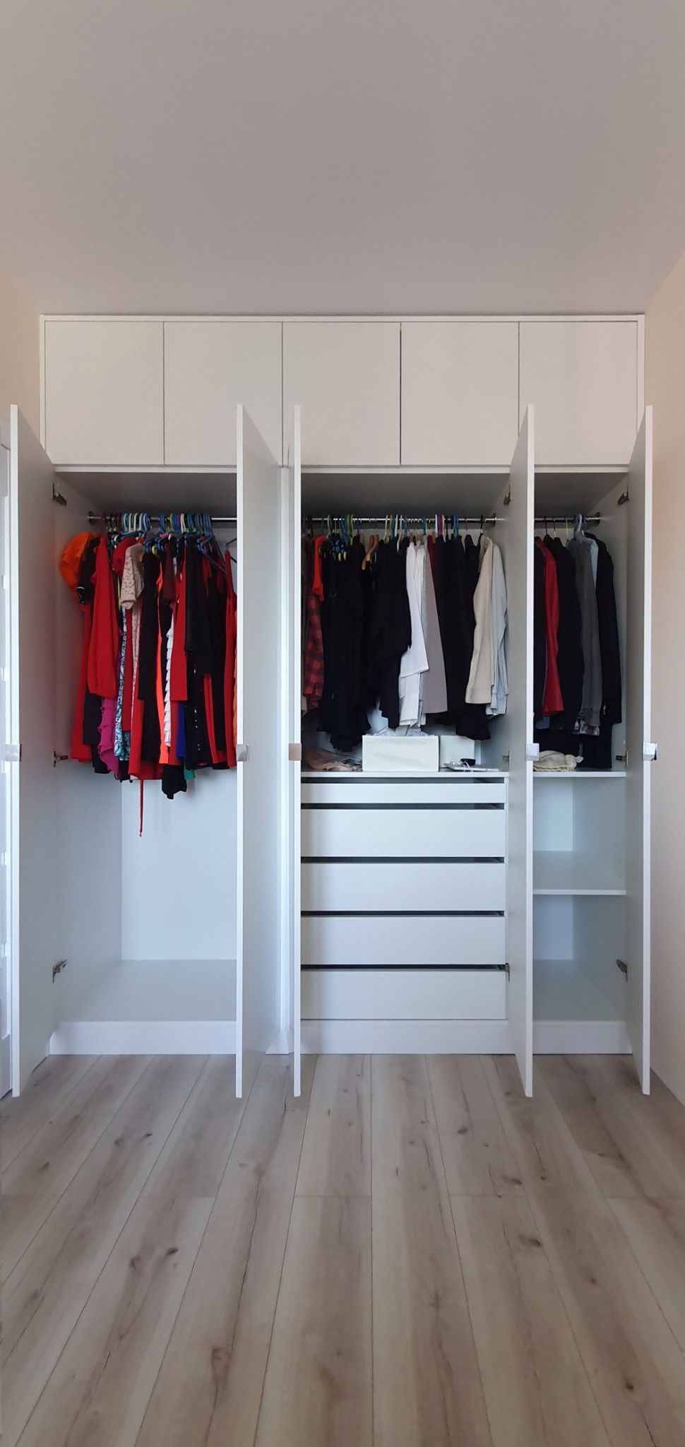 Biała szafa wnękowa z otwartymi drzwiami, widoczne ubrania na wieszakach i szuflady w środkowej części, jasna podłoga z paneli.
