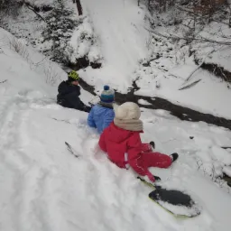 Troje dzieci w zimowych ubraniach siedzi na śniegu obok strumienia, jedno z nich na czarnej desce do zjeżdżania, w tle zasypany śniegiem las.