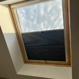 Ciemnoszara, plisowana przesłona okienna w oknie dachowym z widokiem na pochmurne niebo.