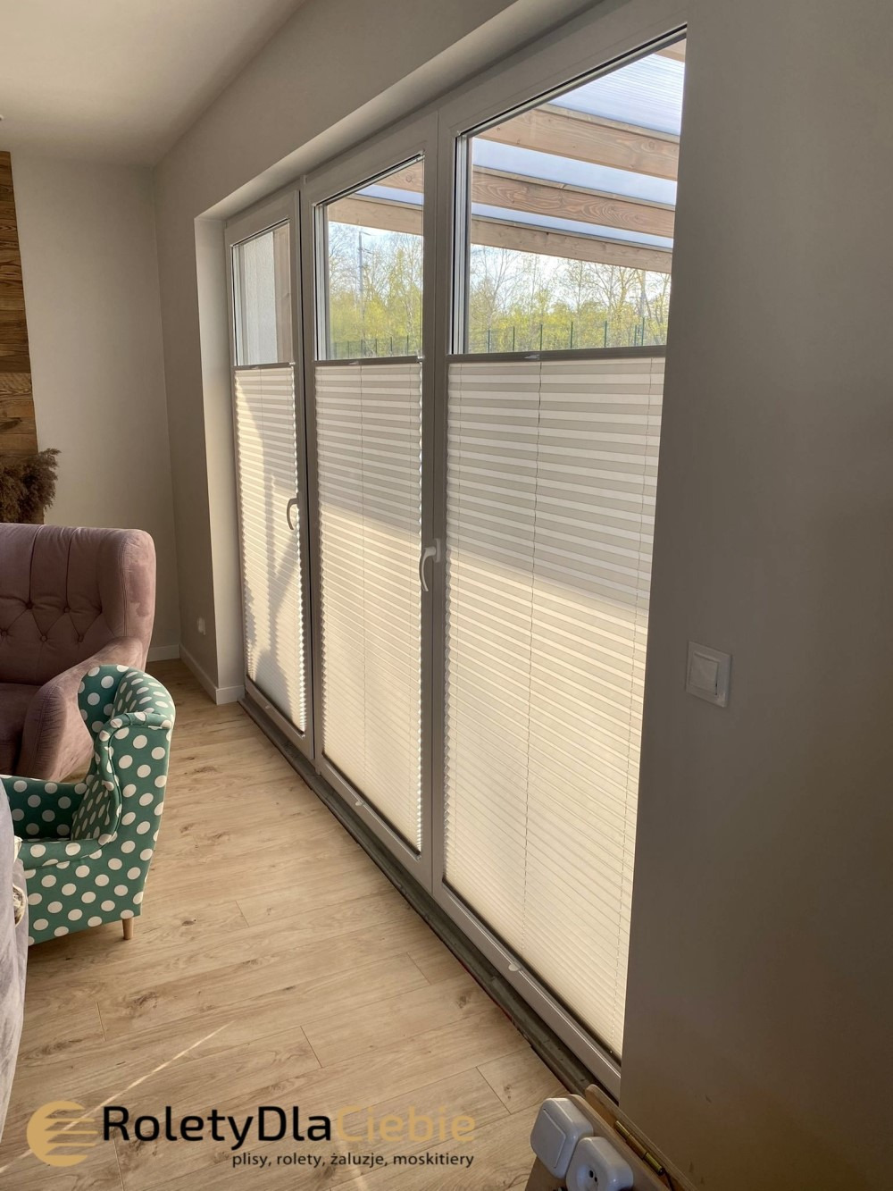 Trzyczęściowe okno balkonowe z zamontowanymi plisami w kolorze kremowym, widok z wnętrza salonu z fotelami w tle.
