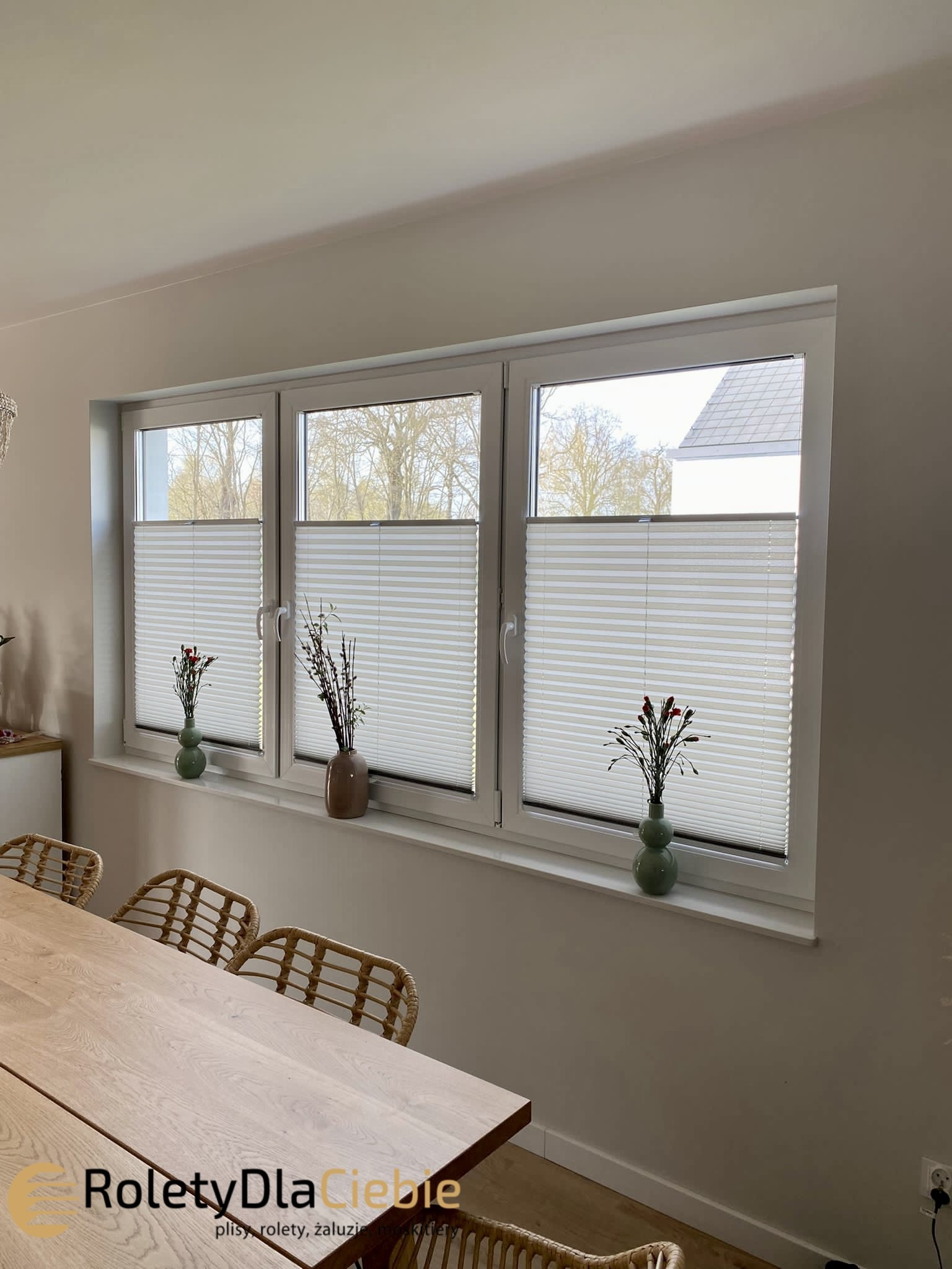 Trzy okna z białymi ramami, osłonięte plisowanymi zasłonami, na parapecie wazony z kwiatami, w tle drzewa bez liści i fragment dachu, w dolnej części widoczny drewniany stół i krzesła z rattanu.