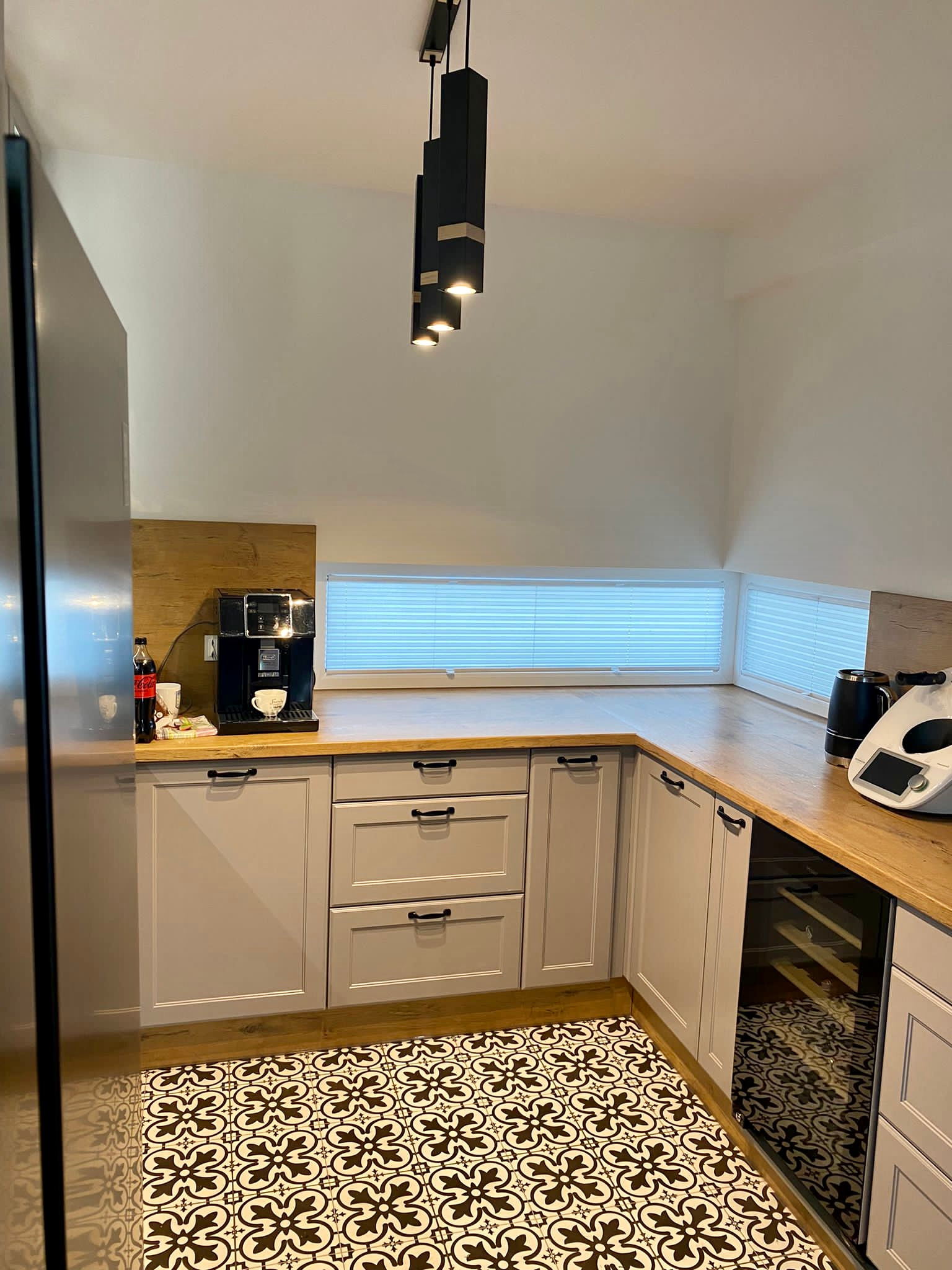 Jasna kuchnia w stylu retro z drewnianym blatem, czarną armaturą i wzorzystą podłogą, okno przesłonięte materiałowymi osłonami okiennymi typu plaster miodu.