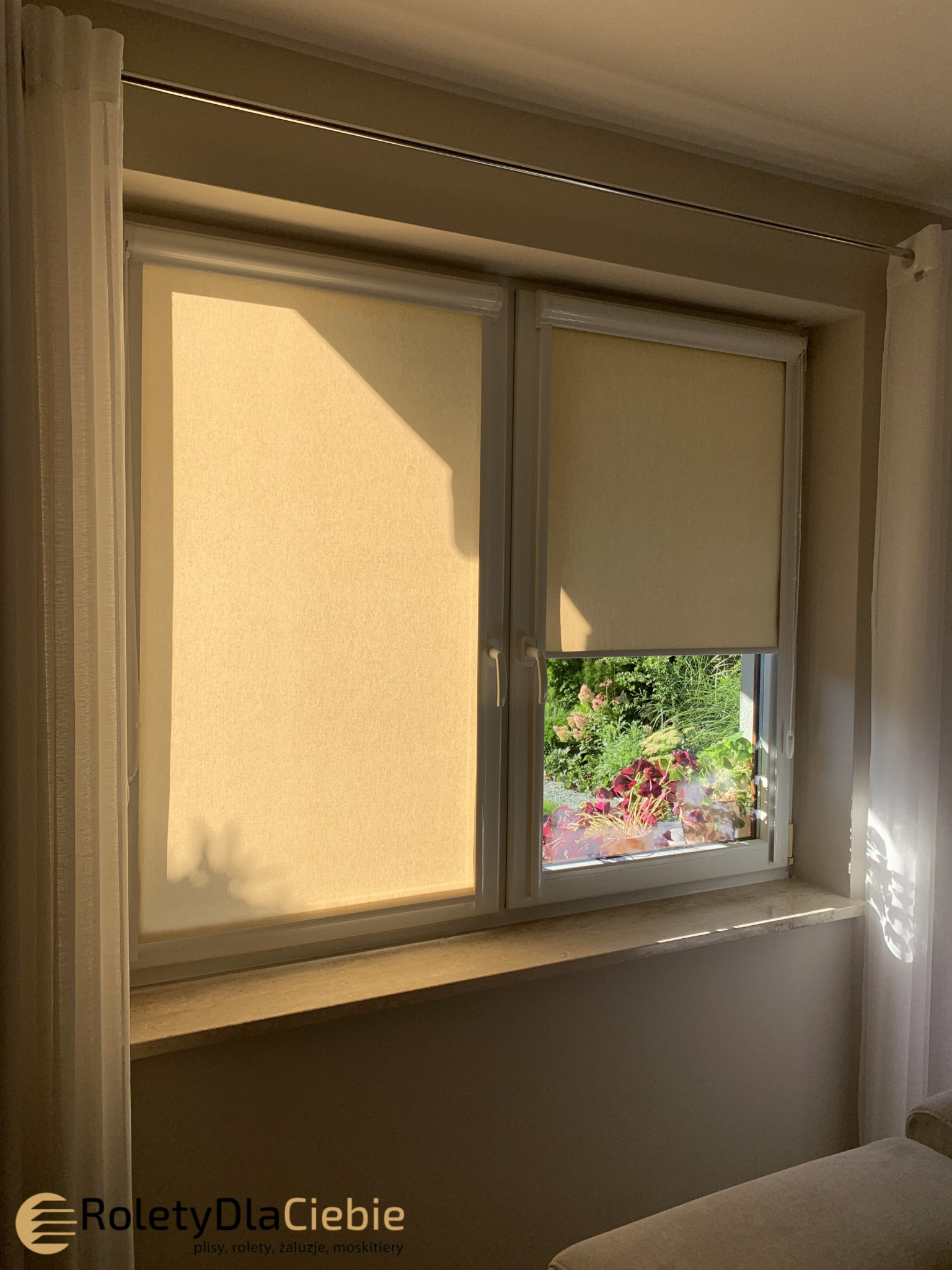 Dwuokienne okno z beżowymi, tekstylnymi osłonami przeciwsłonecznymi; na parapecie doniczki z czerwonymi kwiatami, widok na zielony ogród.