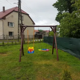 Drewniana huśtawka ogrodowa z dwoma siedziskami: kubełkowym, kolorowym i okrągłym, tęczowym, na tle trawnika, domu z czerwonym dachem i zielonego ogrodzenia.