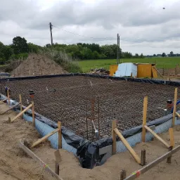 Przygotowany zbrojony fundament pod budowę domu jednorodzinnego, widoczna izolacja pionowa i szalunki, w tle sterta piasku i zabudowania gospodarcze.