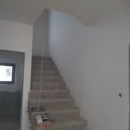 Surowe betonowe schody wewnętrzne w trakcie prac wykończeniowych, widoczne narzędzia i materiały budowlane, świeżo pomalowane ściany na biało, okno w tle.