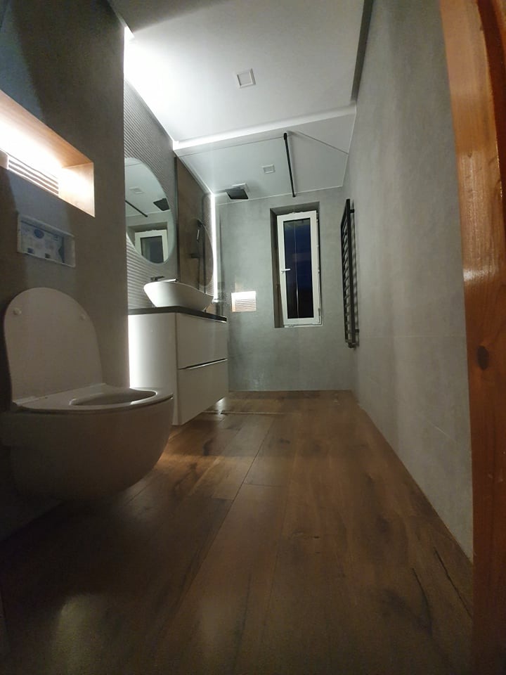 Nowoczesna łazienka z drewnianą podłogą, umywalką nablatową na szafce, owalnym lustrem i minimalistycznym grzejnikiem drabinkowym.