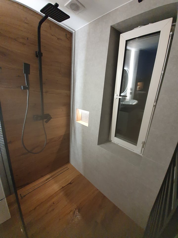 Nowoczesna kabina prysznicowa z drewnopodobną okładziną ścian i podłogi, czarna armatura, odpływ liniowy w podłodze, widok na okno uchylne z widokiem na umywalkę.