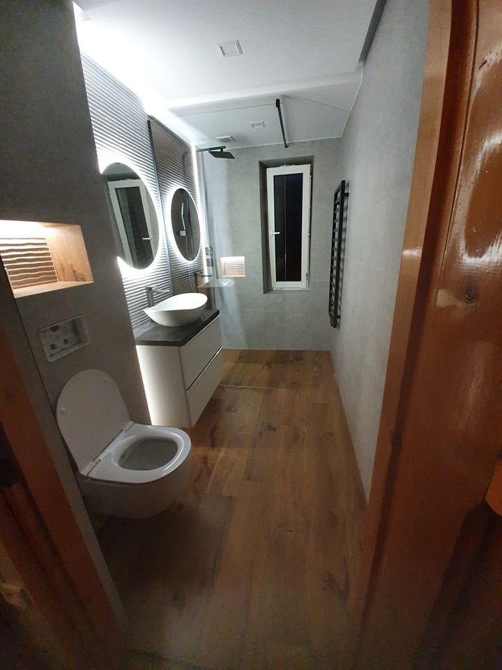 Nowoczesna łazienka z drewnianą podłogą, owalną umywalką nablatową, podświetlanymi okrągłymi lustrami i grzejnikiem drabinkowym. Widok zza otwartych drzwi.