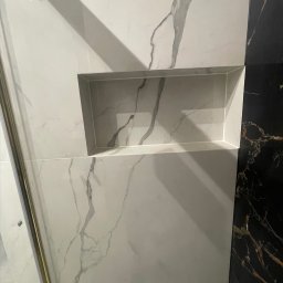 CREO JAKUB CAŁA - Ściana wyłożona białymi płytkami imitującymi marmur z wnęką, obok fragment ściany z czarnymi płytkami z marmurkowym wzorem.