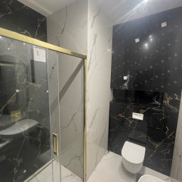 CREO JAKUB CAŁA - Nowoczesna łazienka z kabiną prysznicową o złotych profilach, marmurowymi płytkami w odcieniach czerni i bieli, wiszącą toaletą i umywalką nablatową.