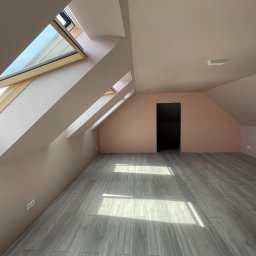 CREO JAKUB CAŁA - Poddasze po remoncie: pomalowane na różowo ściany, szare panele podłogowe imitujące drewno, okna dachowe w drewnianych ramach i wpadające przez nie promienie słoneczne.