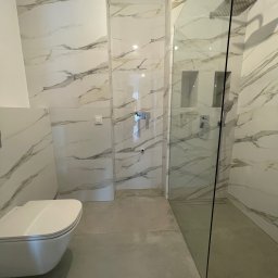 CREO JAKUB CAŁA - Nowoczesna łazienka z białymi płytkami imitującymi marmur, kabiną prysznicową typu walk-in z czarnymi elementami, podtynkową baterią, wiszącą toaletą i szarą podłogą.
