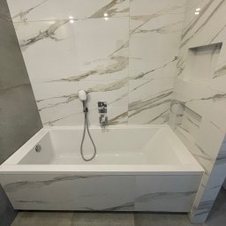 CREO JAKUB CAŁA - Nowoczesna łazienka z białą wanną obudowaną marmurkowymi płytkami, szara ściana z płytek po lewej stronie, wnęka w ścianie nad wanną.