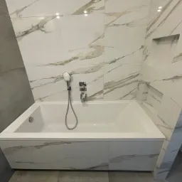 Nowoczesna łazienka z białą wanną obudowaną marmurkowymi płytkami, szara ściana z płytek po lewej stronie, wnęka w ścianie nad wanną.