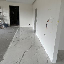 CREO JAKUB CAŁA - Układanie płytek imitujących marmur w nowym mieszkaniu, widoczne przygotowane ściany i surowa posadzka w sąsiednim pomieszczeniu, drabina i narzędzia w tle.