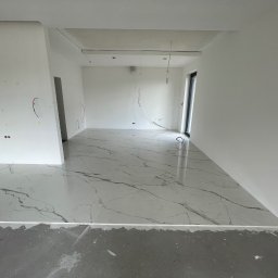 CREO JAKUB CAŁA - Nowoczesne wnętrze w trakcie prac wykończeniowych: białe ściany z widocznymi przewodami elektrycznymi, marmurowa podłoga z szarymi żyłami i surowa wylewka przy ścianach.